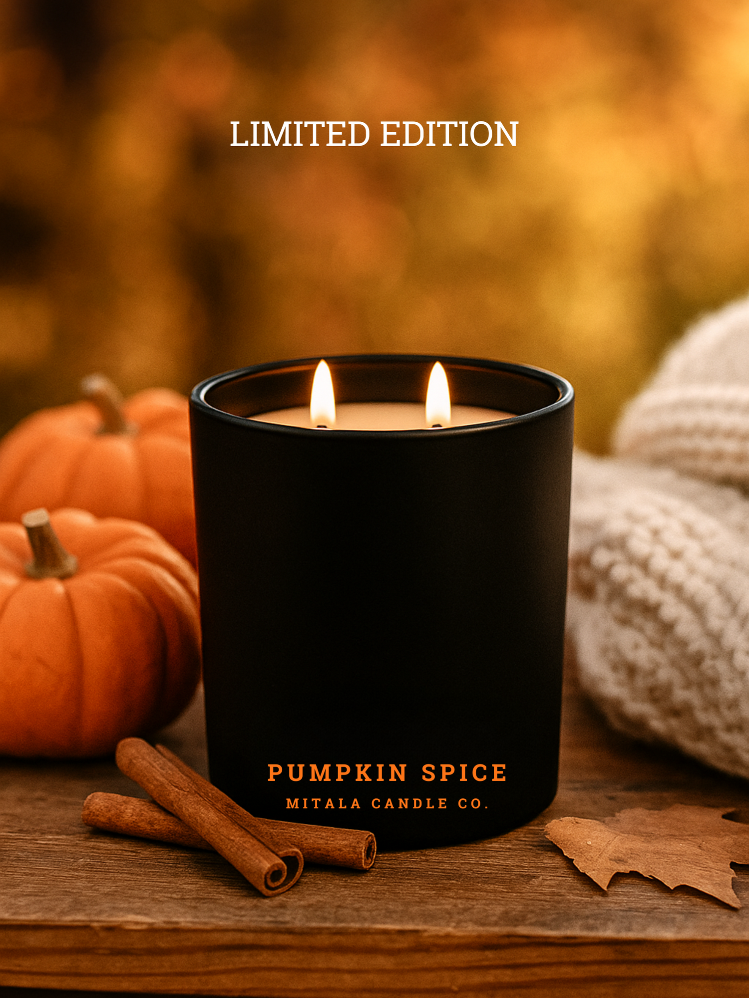 A Mitala Candle Co. candle labeled "Pumpkin Spice" with two flames burns on a wooden table, surrounded by small pumpkins, cinnamon sticks, and a cozy white blanket. Warm autumn tones.