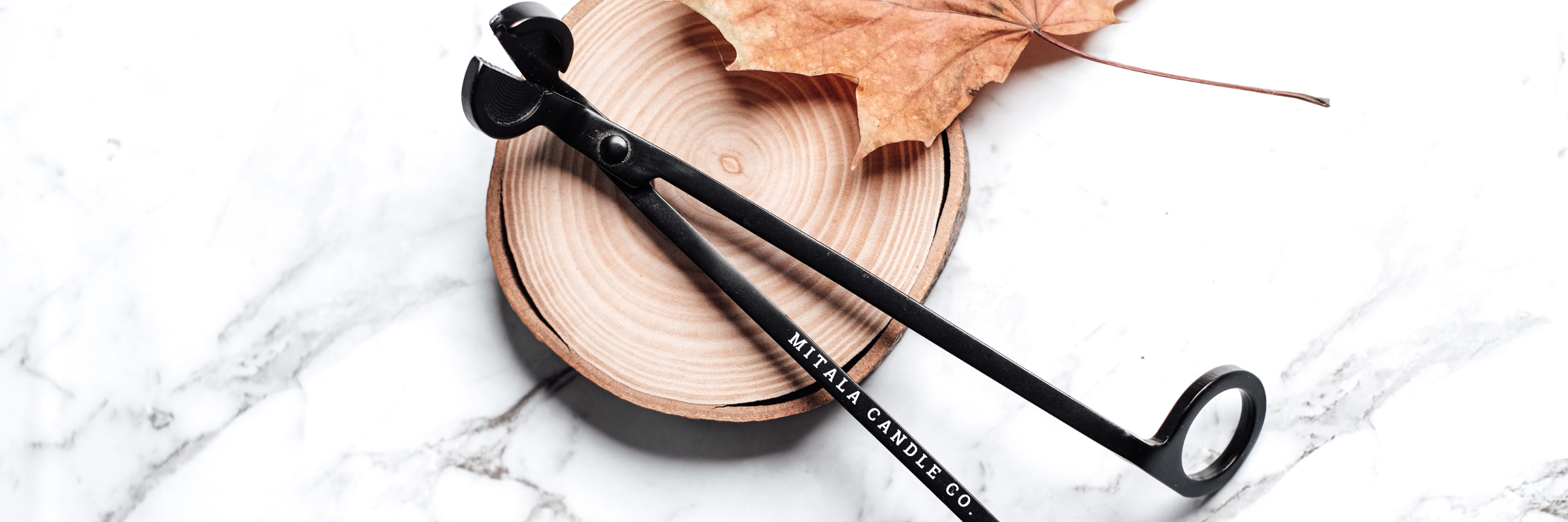 Black candle wick trimmer resting on a round wooden coaster, with a dried brown leaf nearby, all placed on a white marble surface.