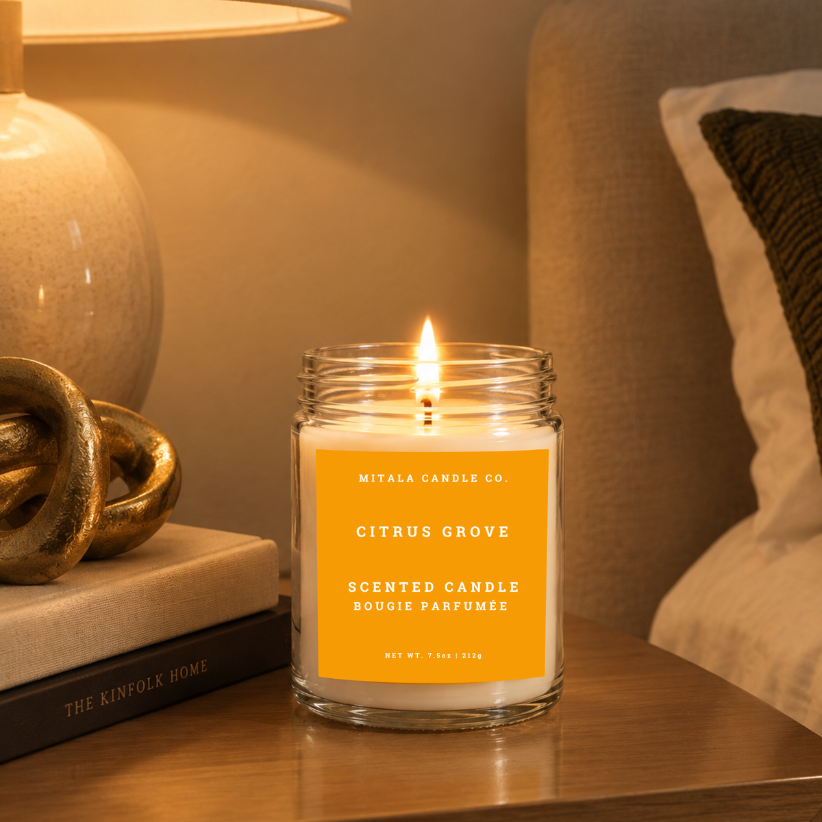A 7.5 oz scented candle in a clear glass jar with a white wax fill and a prominent orange label reading 'Mitala Candle Co. Citrus Grove Scented Candle' sits on a wooden bedside table beside a lamp and decor, creating a warm, cozy mood.