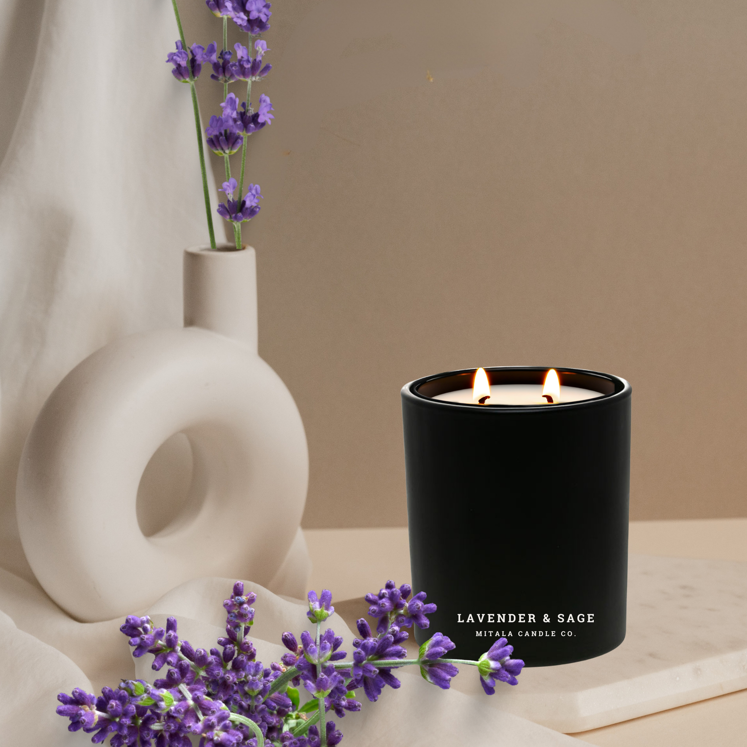 A black Mitala candle labeled "Lavender & Sage" with two lit wicks sits on a beige surface, surrounded by purple lavender flowers and a ceramic vase.