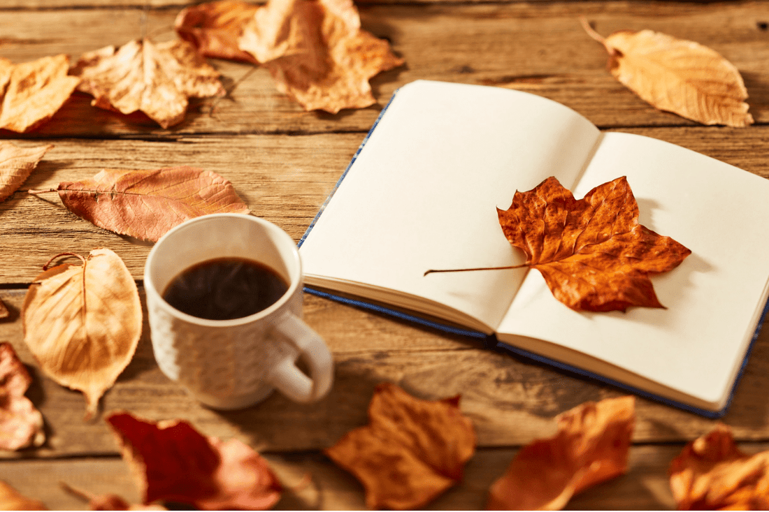 A cup of coffee, an open journal, and autumn leaves scattered on a rustic wood table, the perfect cozy fall setting.