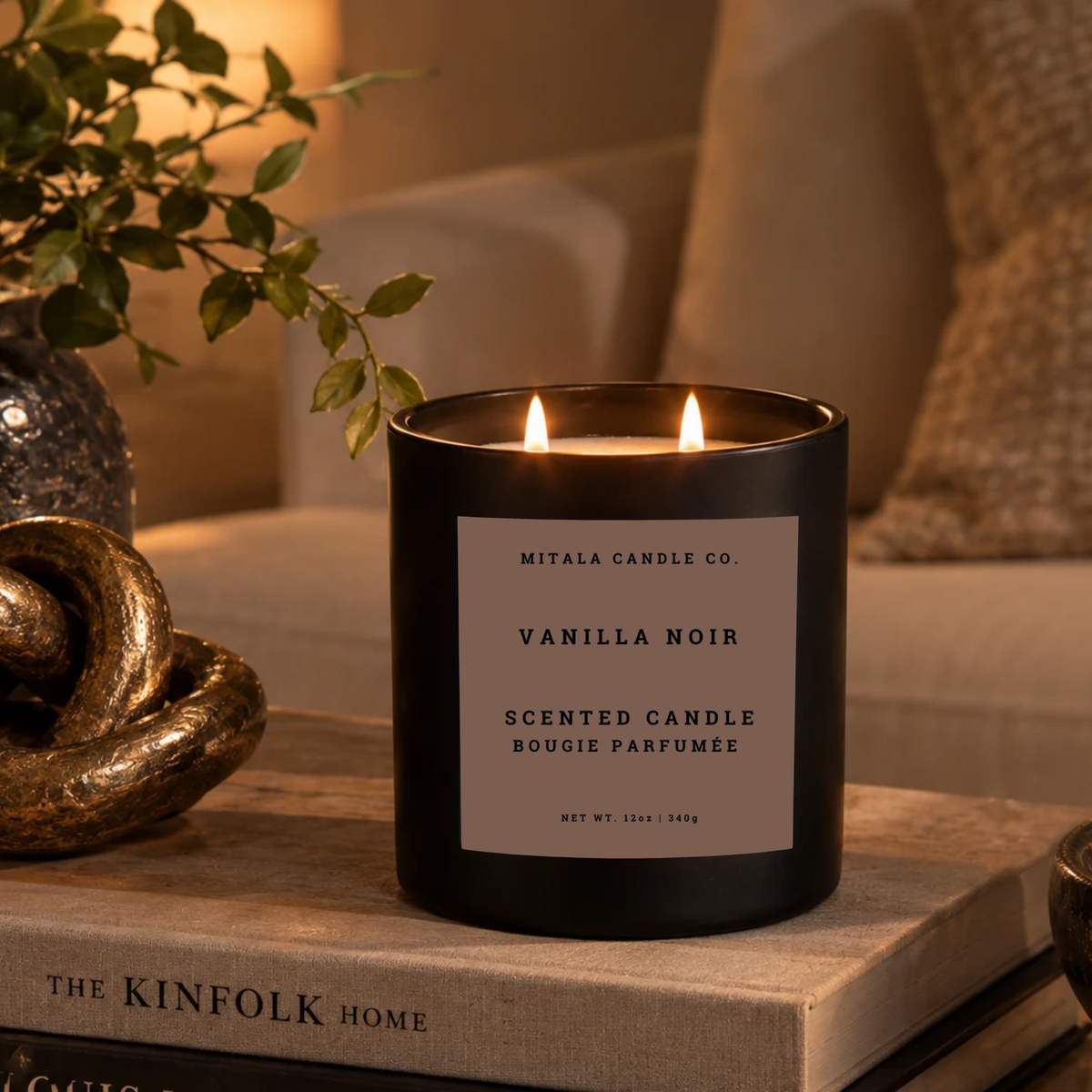 A black scented candle jar with two lit flames sits on a book in a warm, styled indoor setting. The front label reads 'MITALA CANDLE CO. Vanilla Noir Scented Candle',  beside leafy branches in a warm, tranquil living room.