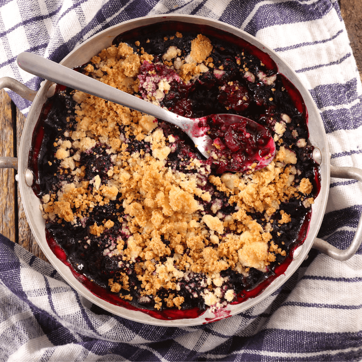 Blueberry crumble in round baking dish on striped towel with spoon showing berry filling and golden crumb topping