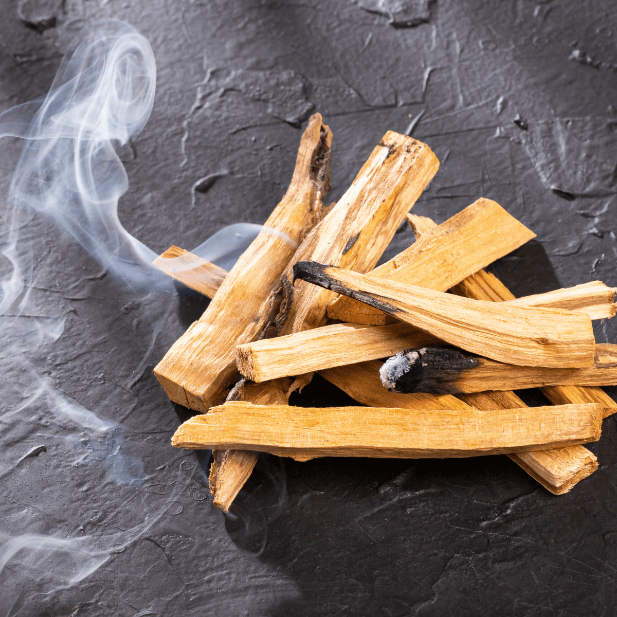 Pile of palo santo sticks emitting fragrant smoke against textured black background, swirling gently to create a calming atmosphere
