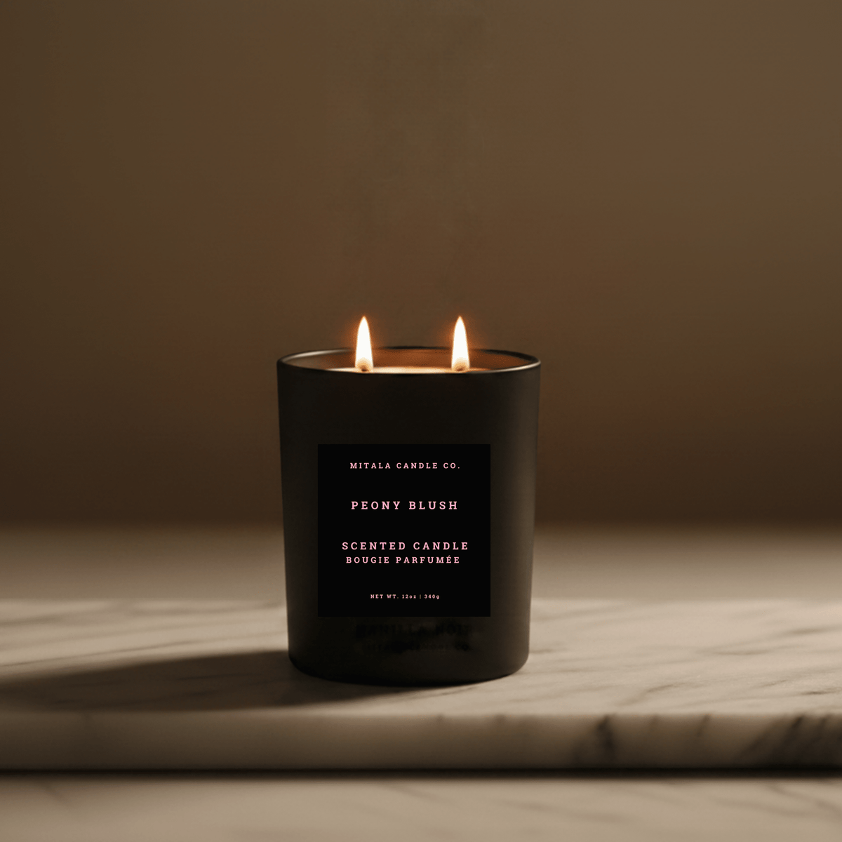 A black double-wick scented candle labeled 'Mitala Candle Co. Peony Blush' with two lit flames, set on a light marble surface against a warm brown background.