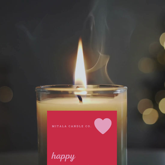 A lit flickering candle with gentle smoke wafting up, set against a dark background. A red overlay reads "Mitala Candle Co." and "happy Valentine's Day", with a pink heart icon. Calm ambiance.