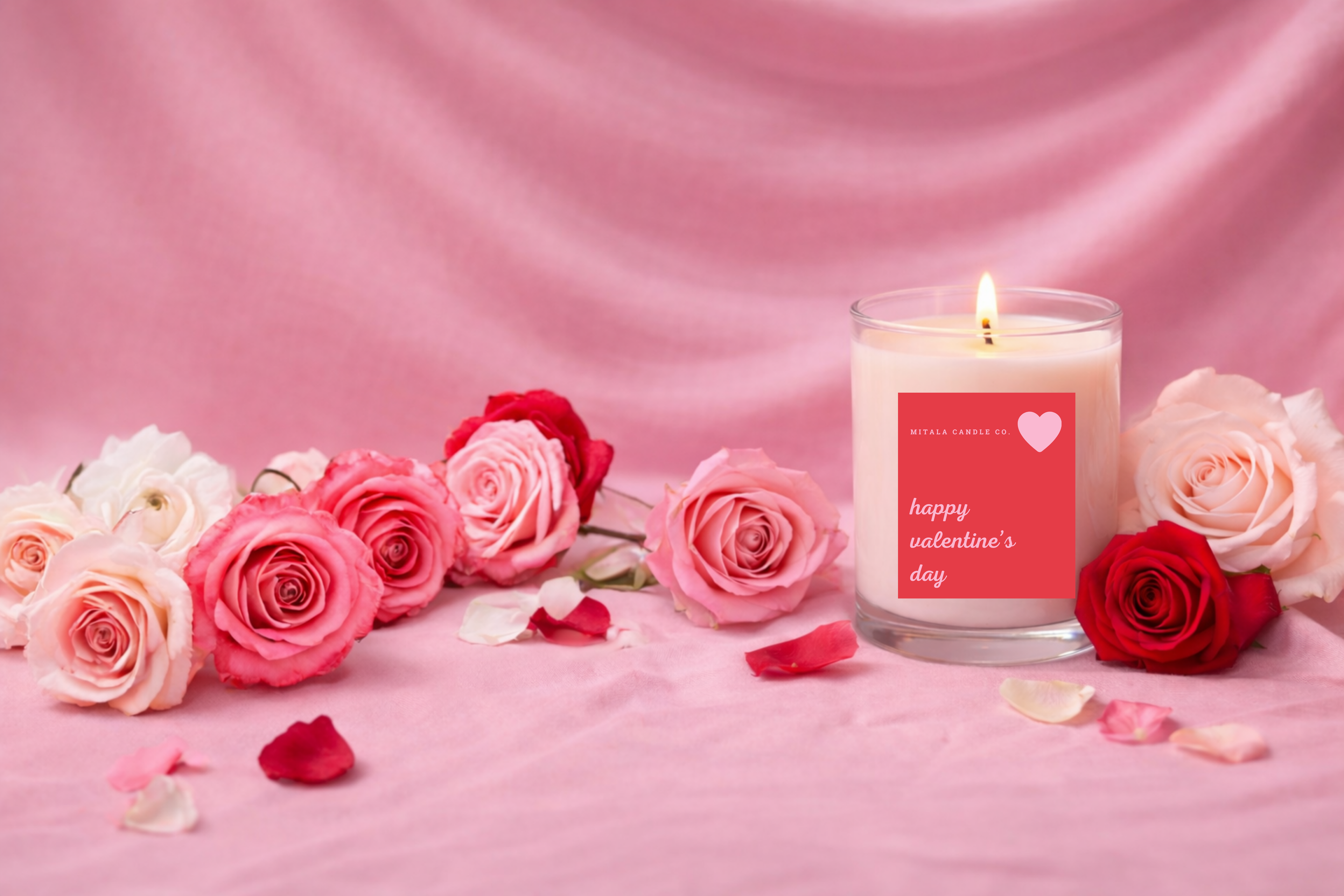 A lit Mitala Candle Co.  candle in a glass jar with "Happy Valentine's Day" label, surrounded by pink and red roses on a soft pink fabric, creating a romantic feel.
