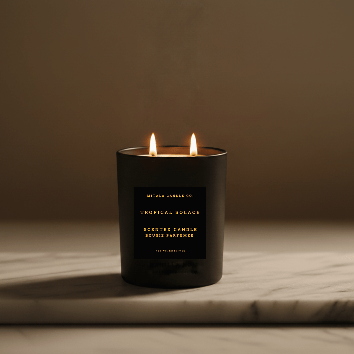 A black double-wick scented candle labeled 'Tropical Solace' by Mitala Candle Co., lit with two flames, set on a light marble surface against a warm brown background.