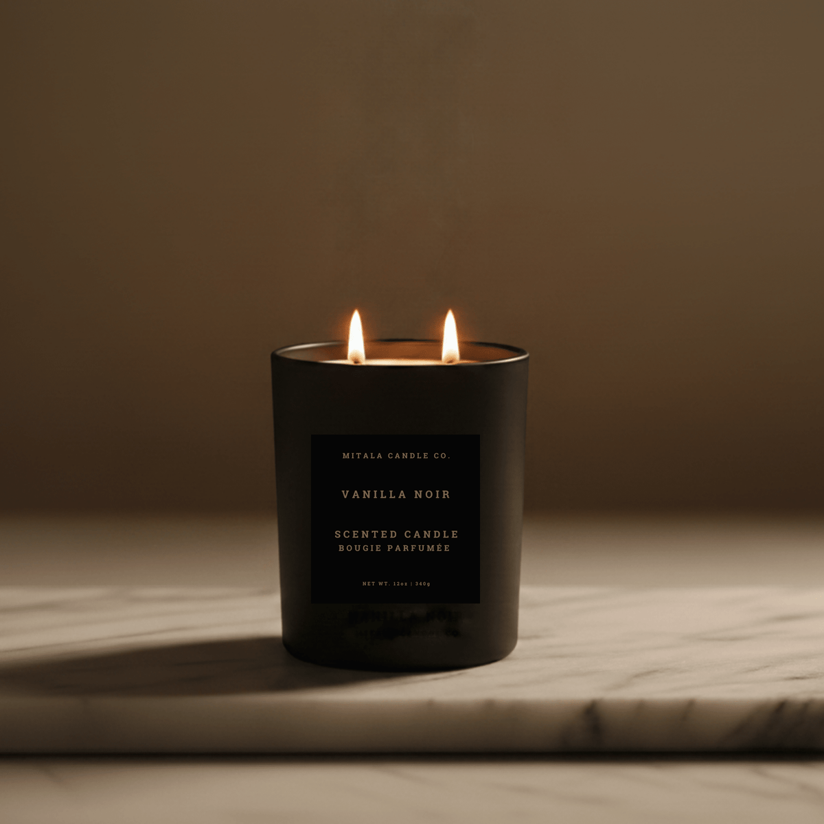 A black double-wick scented candle labeled 'Mitala Candle Co. Vanilla Noir' with two lit flames, set on a light marble surface against a warm brown background.